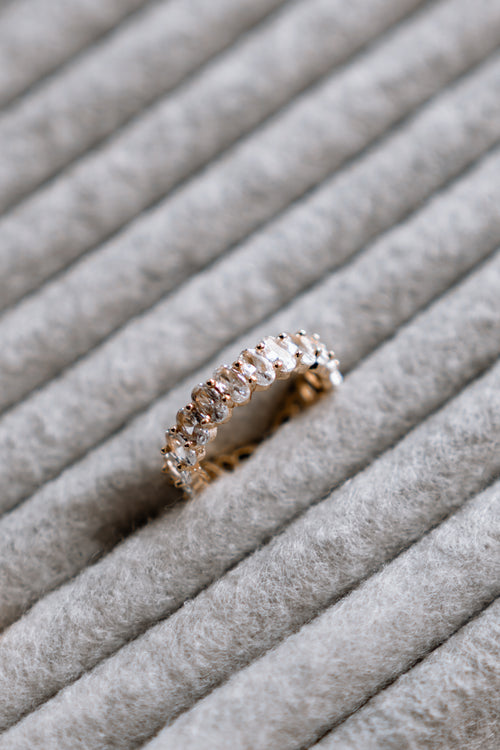 A gold eternity ring with a row of clear round-cut gemstones, resting on a textured gray felt ring stand.