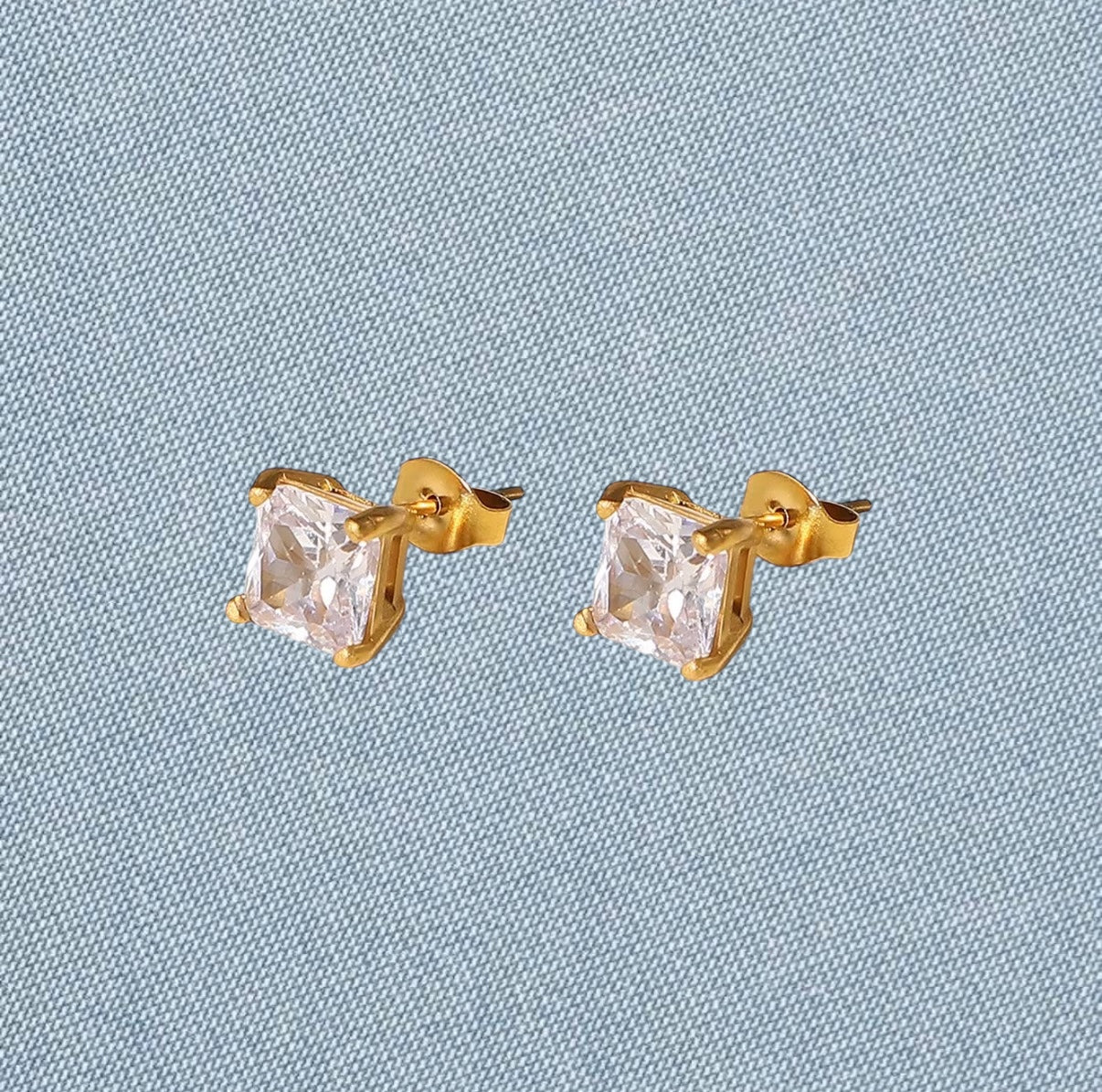 Gold stud earrings with clear gemstones on a light blue textured background